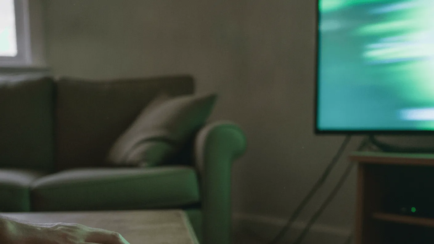 A hand reaches for a remote control on a wooden coffee table, with a glowing green TV screen and sofa in the background.